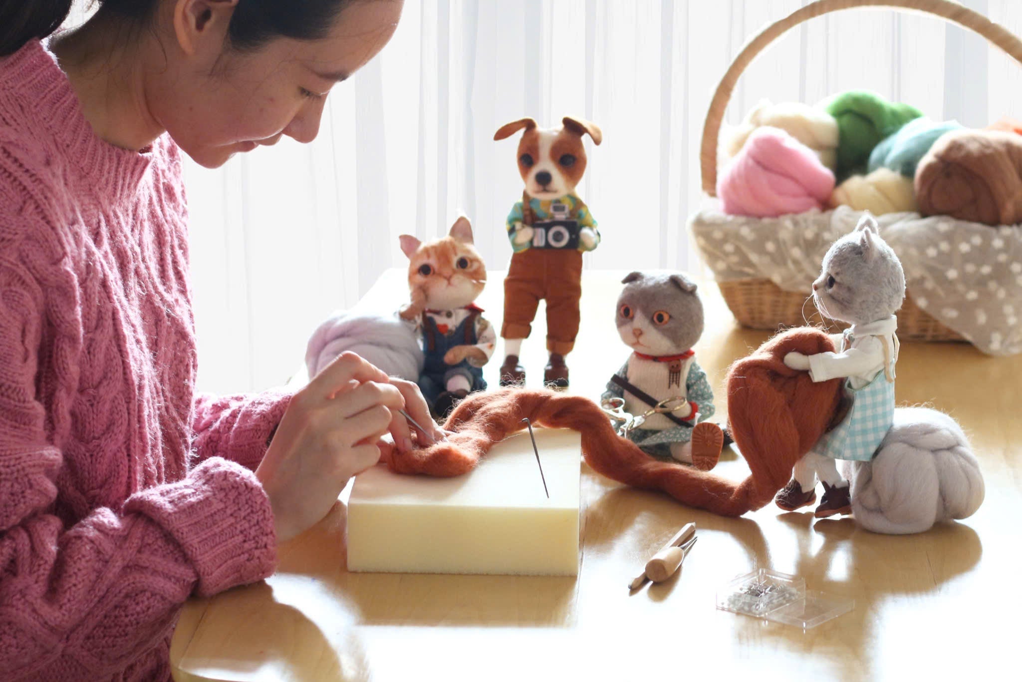 Woman in pink sweater working on a needle felted project with handmade animal figures and wool at a table.